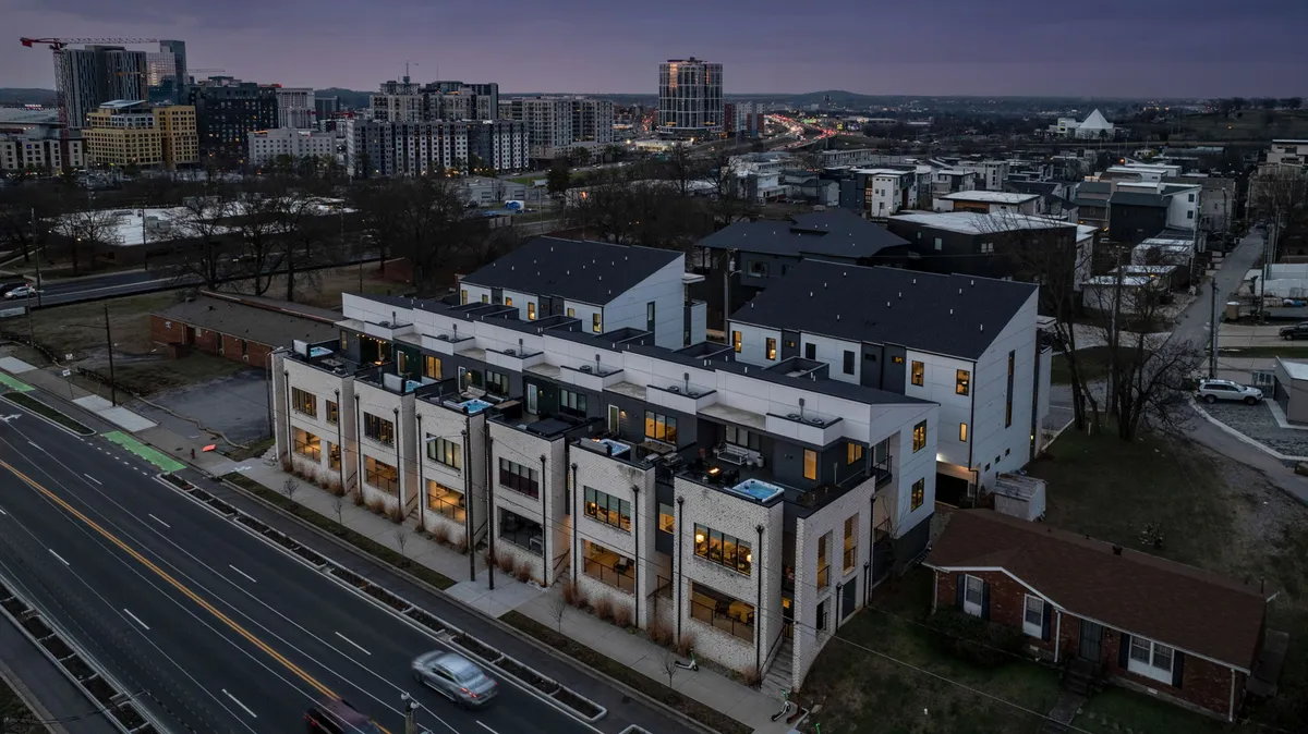 This exterior image captures a row of modern townhouse architecture set against an urban skyline, with visible street activity. People can be seen walking and socializing, and the scene is vibrant and contemporary, reflecting the lively energy of a developing district.