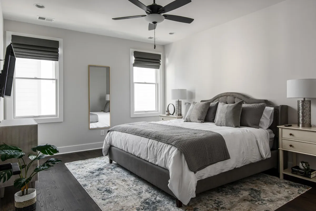 A serene bedroom featuring a king-sized bed dressed in soft gray bedding, a mirrored dresser reflecting ample natural light from large windows with shades, and a plush decorative rug underfoot. The mood is tranquil and restful, making it an ideal retreat for unwinding.
