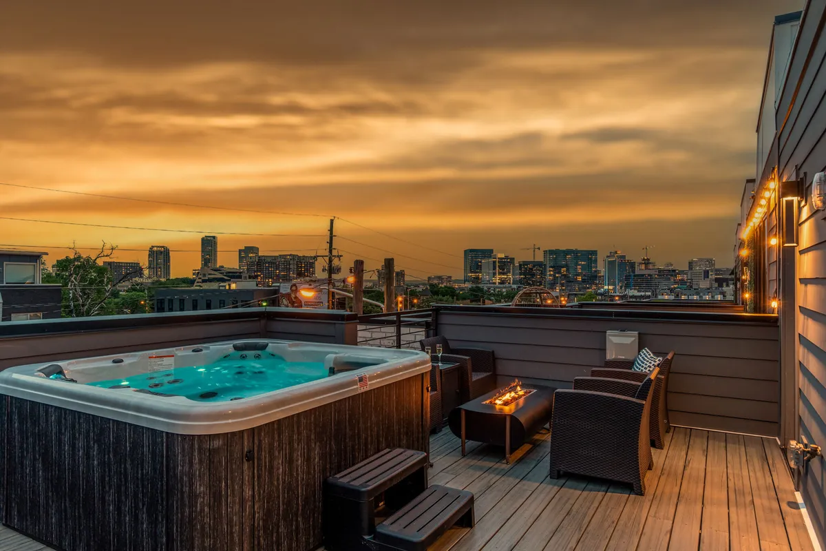 The image features a stunning outdoor space with a luxurious hot tub, elegant seating area, and twinkling decorative lights, all set against the backdrop of Nashville’s city skyline at sunset. The ambiance is both serene and sophisticated, inviting guests to unwind, soak in the view, and share memorable moments with friends or family.