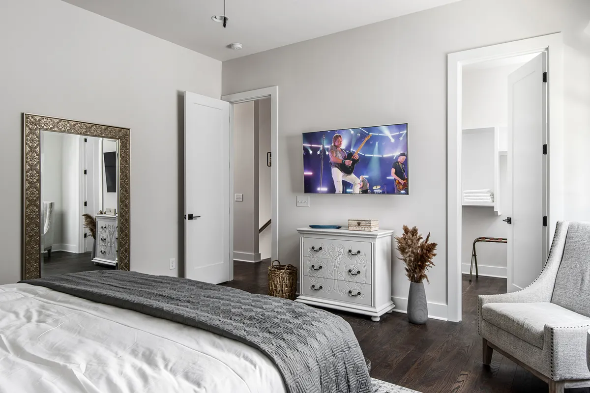 This image depicts a serene and inviting bedroom featuring a queen-size bed draped with a textured blanket, a large mirror, a TV displaying a concert scene, a decorative plant in a vase, and a nightstand adorned with decorative items. The overall feel is one of comfort and privacy, with modern touches and thoughtful amenities.