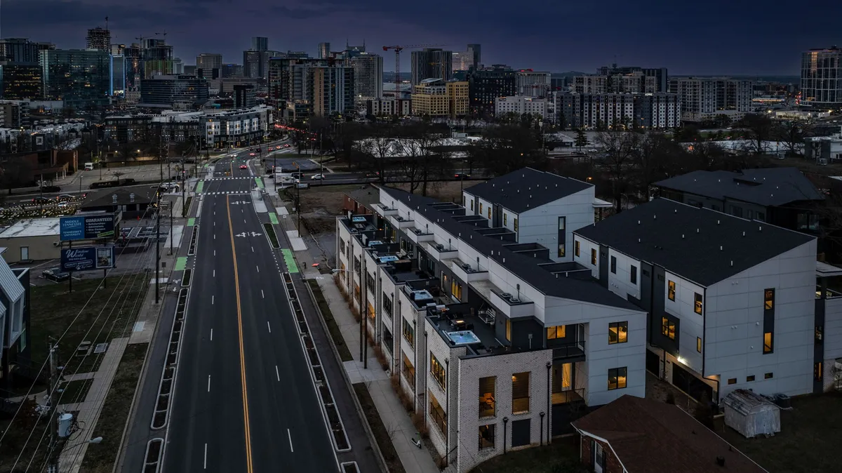 A vibrant exterior view of modern townhouses set against a glowing city skyline at dusk. Rooftop spaces are visible, and city lights start to shimmer, evoking an atmosphere of lively urban adventure and evening family gatherings.