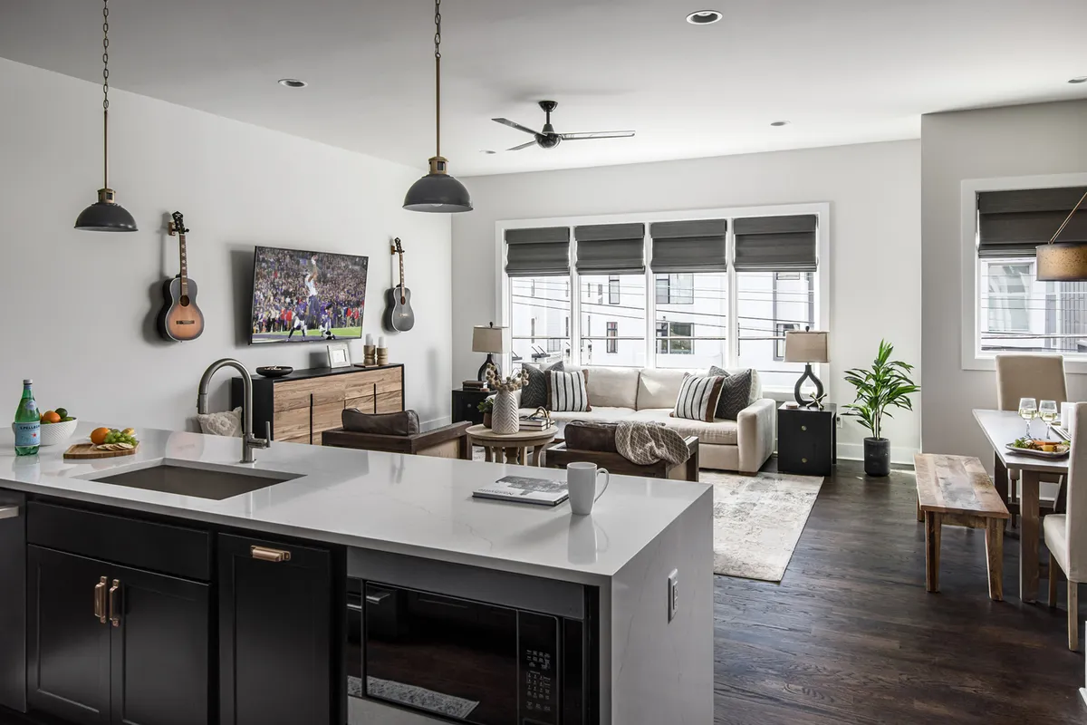 An open-concept living, dining, and kitchen area featuring a kitchen island with a sink, a television displaying a sports event, a wall-mounted guitar, a comfortable sofa with decorative cushions, and a dining table set for a meal. The layout is airy and conducive to both lively gatherings and relaxed moments.