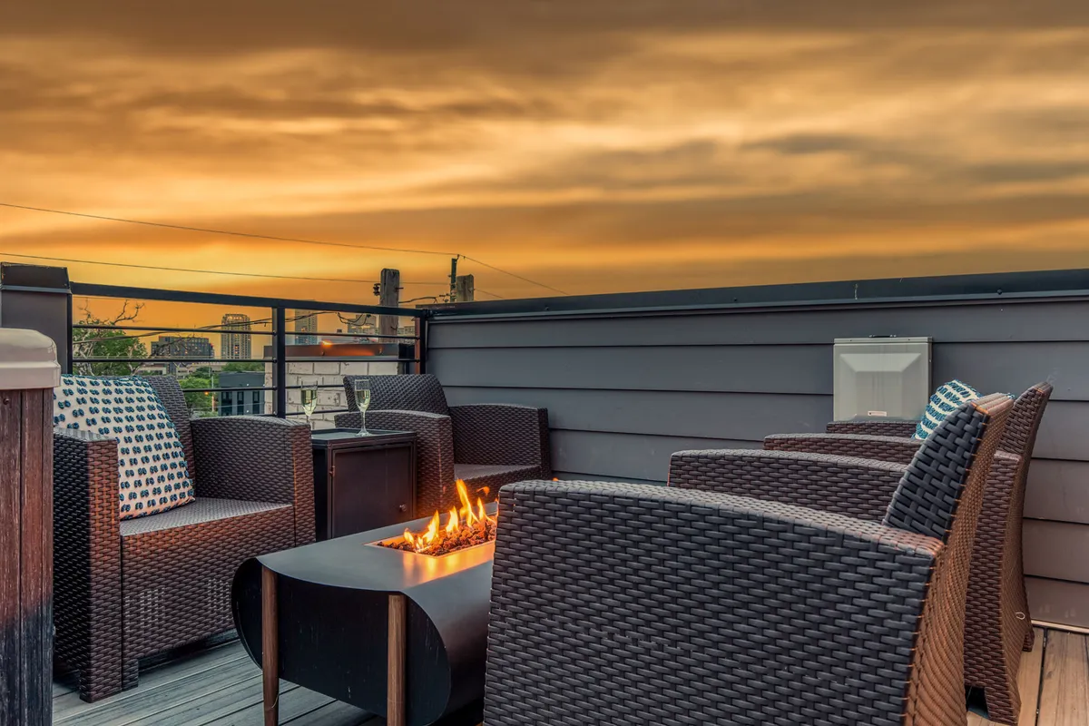 An inviting rooftop space with a flickering fire pit, plush outdoor sofas, and a brilliant sunset sky overhead. The scene is alive with the potential for lively or laid-back gatherings, blending comfort and urban sophistication.