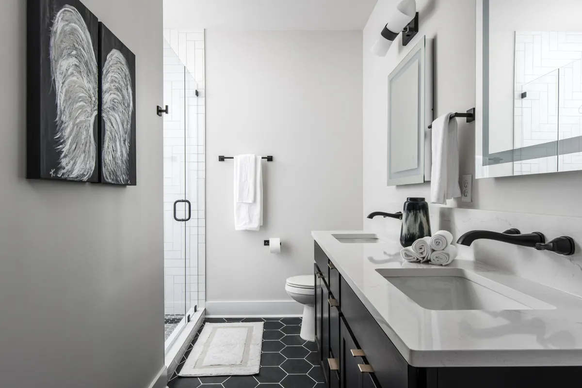 A sophisticated bathroom with a double vanity featuring sleek countertops, a spacious glass shower enclosure, eye-catching hexagonal floor tiles, and striking black-and-white wing artwork on the wall. The atmosphere is refined, clean, and spa-like.